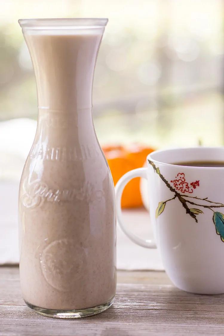 Pumpkin spice creamer in an old fashioned glass milk bottle, next to a cup of coffee, with pumpkins in the background.