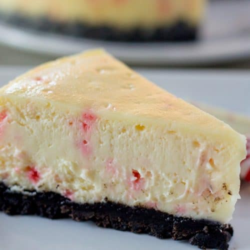 A slice of white chocolate peppermint bark cheesecake on a white plate with a piece of peppermint bark and a starlight mint.