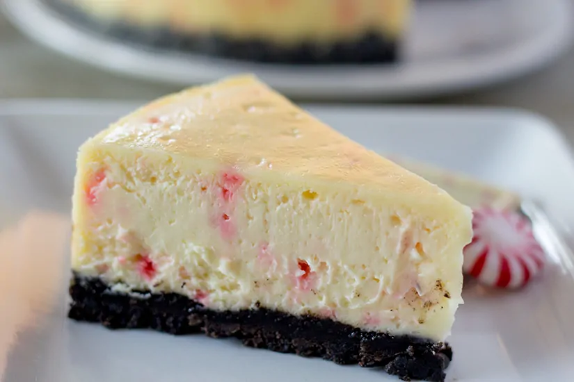 A slice of white chocolate peppermint bark cheesecake on a white plate with a piece of peppermint bark and a starlight mint.