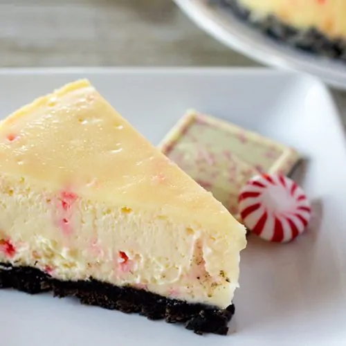 A slice of white chocolate peppermint bark cheesecake on a white plate with a piece of peppermint bark and a starlight mint.