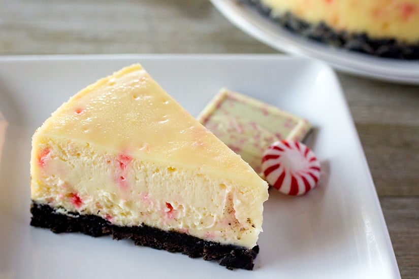 A slice of white chocolate peppermint bark cheesecake on a white plate with a piece of peppermint bark and a starlight mint.