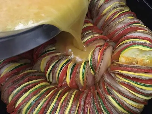 pouring eggs over thinly sliced potatoes, tomatoes, zucchini, and squash in ratatouille pattern