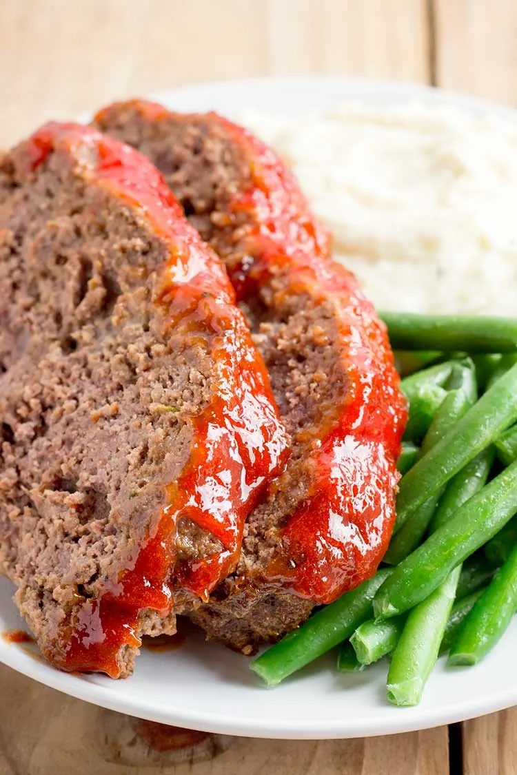 sliced meatloaf on white plate with mashed potatoes and green beans