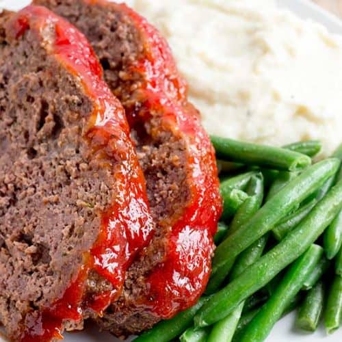 sliced meatloaf on white plate with mashed potatoes and green beans