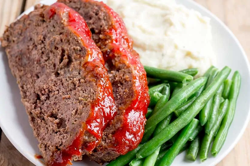 sliced meatloaf on white plate with mashed potatoes and green beans