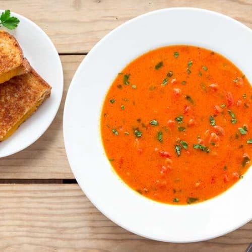Tomato basil soup in white bowl with grilled cheese on side plate.