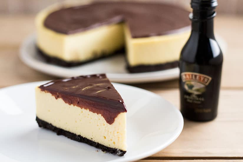 Slice of cheesecake, covered in chocolate sauce, on plate next to a small bottle of Bailey's.