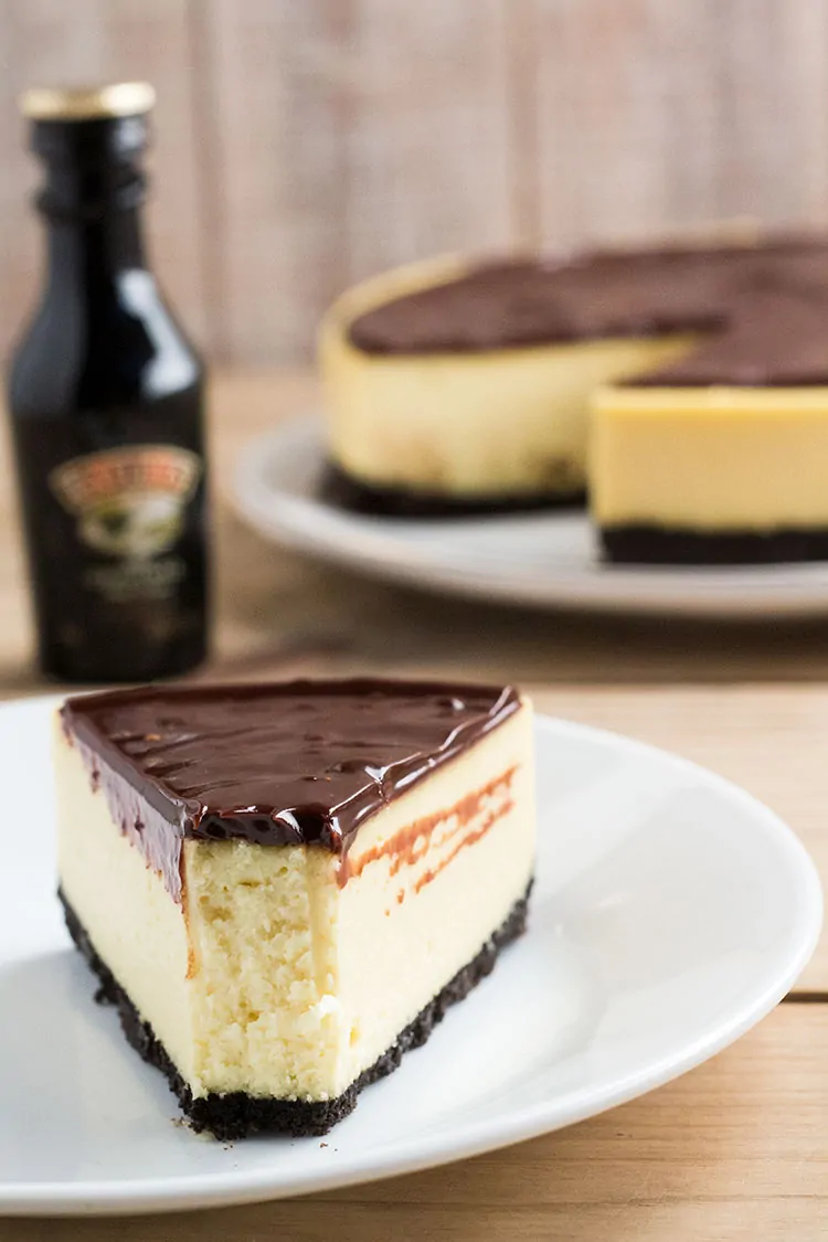 Slice of cheesecake, covered in chocolate sauce, in front of small bottle of Bailey's.