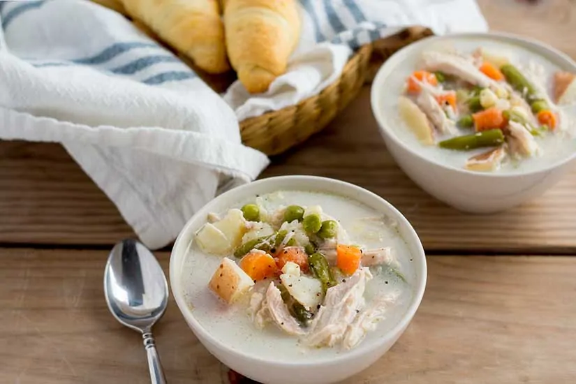 Two bowls of chicken pot pie soup with basket of crescent rolls.