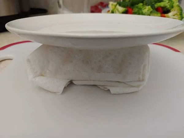 Tofu wrapped in paper towels topped with small plate.