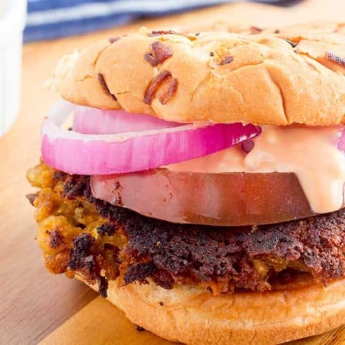 Vegan burger on cutting board with red onions, tomato, and special sauce.