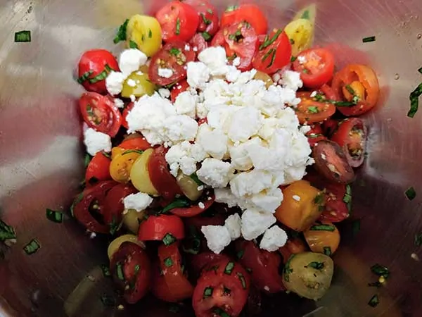 Sliced grape tomatoes, chiffonade basil, and garlic in mixing bowl topped with feta cheese.