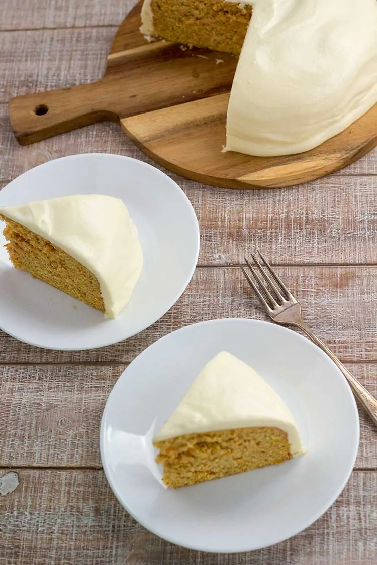 Two slices of carrot cake on whites plate with whole cake in background.