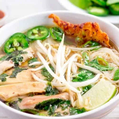 Instant Pot chicken pho in white bowl with garnishes in background.
