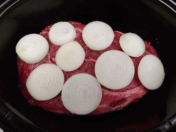 Uncooked chuck roast topped with sliced onions in slow cooker.