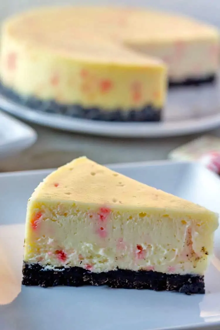A slice of white chocolate peppermint bark cheesecake on a plate with the whole cheesecake in the background.