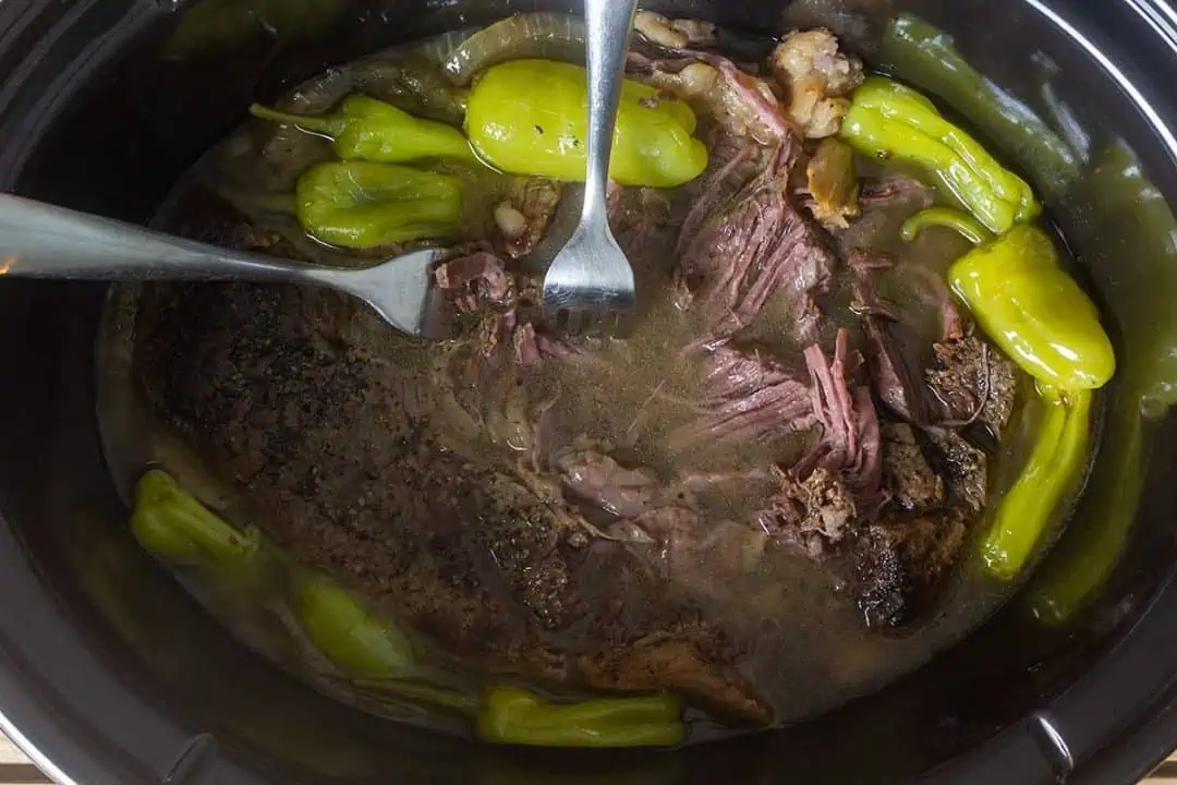 Pot roast in slow cooker with peppers and onions being pulled apart with two forks.
