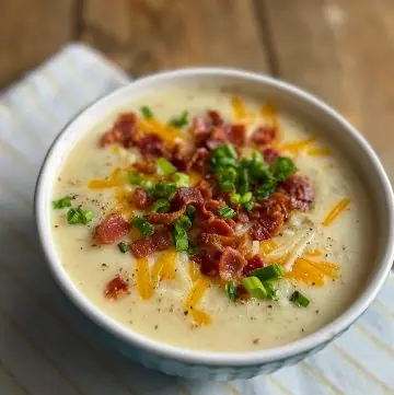 loaded potato soup in a white-rimmed bowl on top of a white and yellow striped linen napkin on a wooded table.