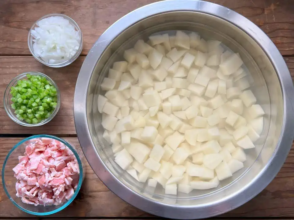 chopped potatoes soaking in water in stainless steel bowl next to three glass bowls with diced onions, diced peppers, and chopped bacon, all on wood tabletop.