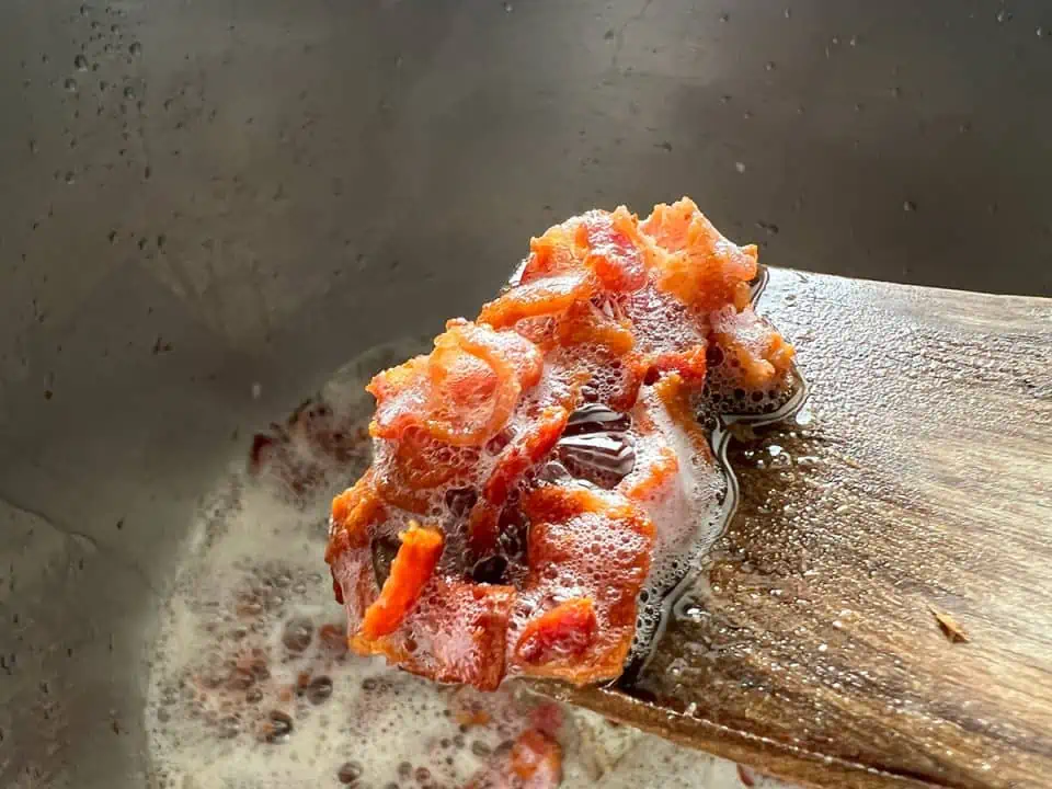 closeup of crispy bacon pieces on a wooden spatula with rendered fat at the bottom of Instant Pot in the background.
