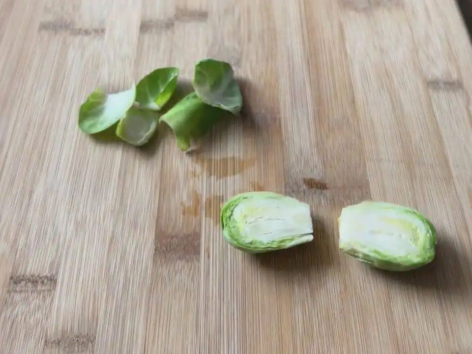 One trimmed Brussels sprout cut in half lengthwise next to the loose leaves on a wooden cutting board.