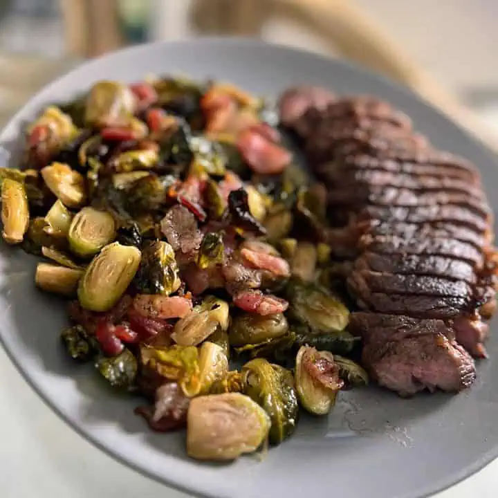 Maple bacon Brussels sprouts on a gray plate with a sliced sirloin steak.