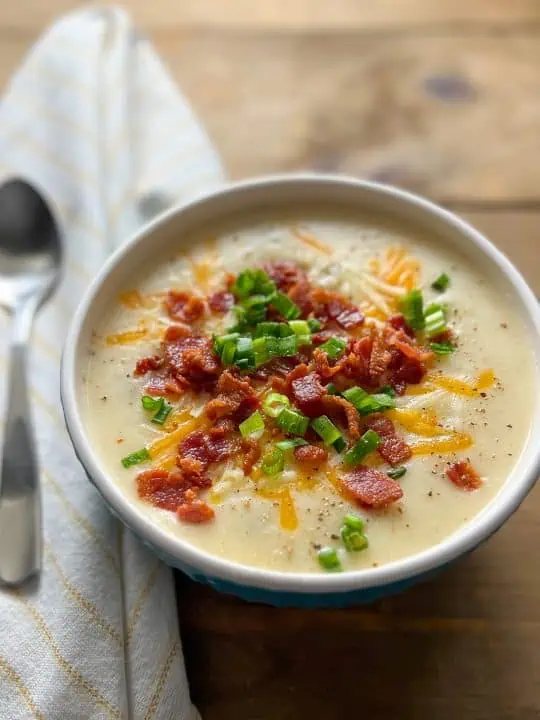 loaded potato soup in a white-rimmed bowl next to a spoon on top of a white and yellow striped linen napkin on a wooded table.