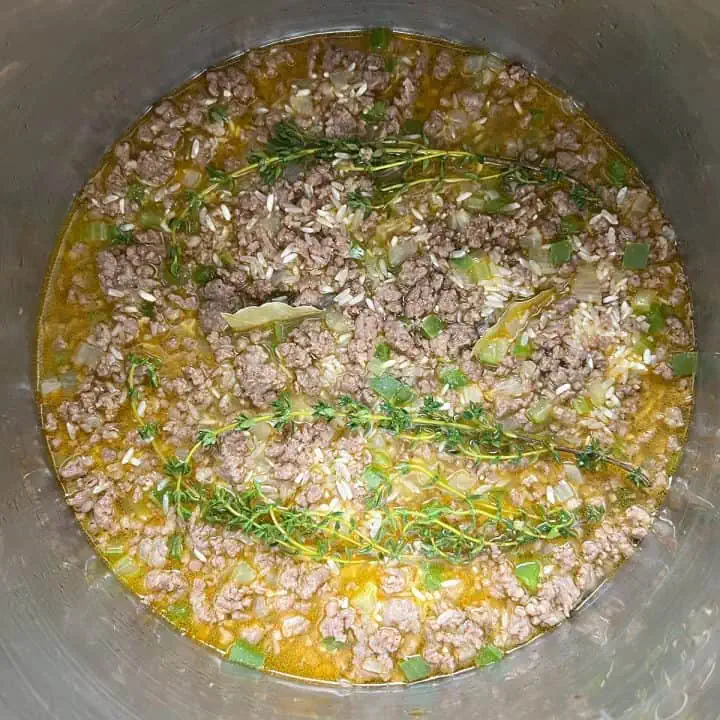 Browned meat and uncooked rice with chicken broth in an Instant Pot topped with thyme sprigs and bay leaves.