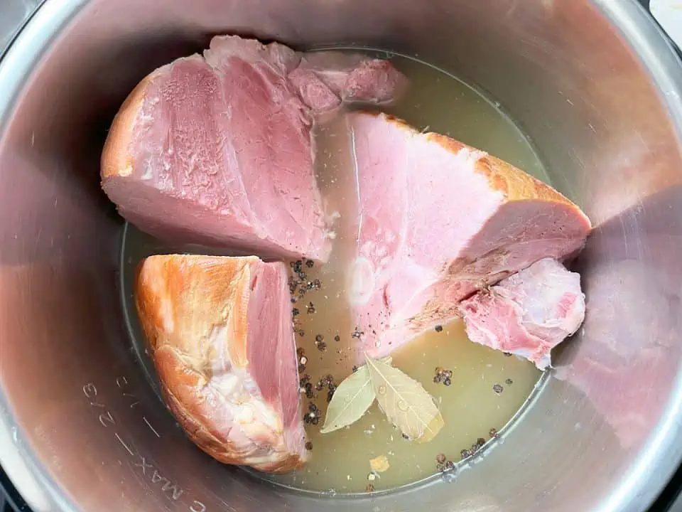 Large chunks of ham in an Instant Pot with broth, bay leaves, and peppercorns.