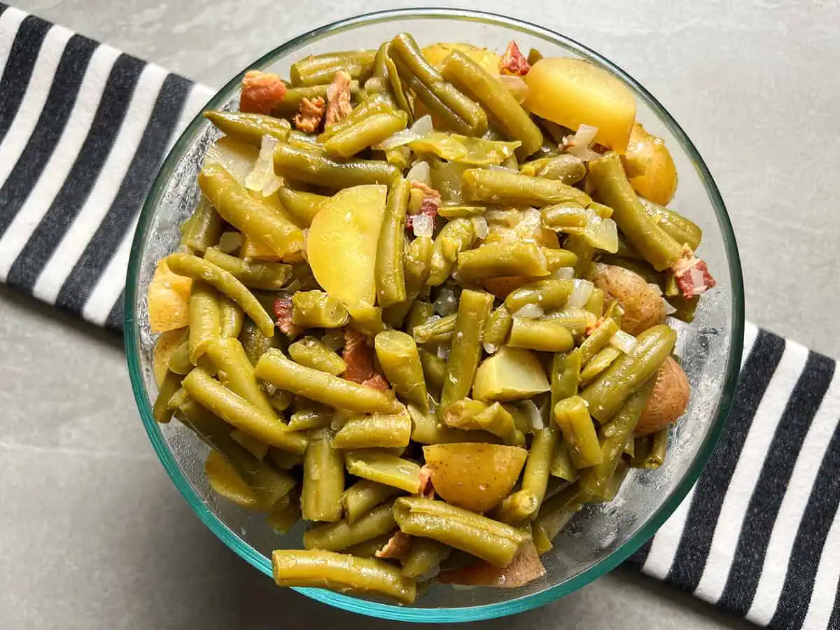 Green beans with bacon and potatoes in glass bowl on top of black and white striped linen.