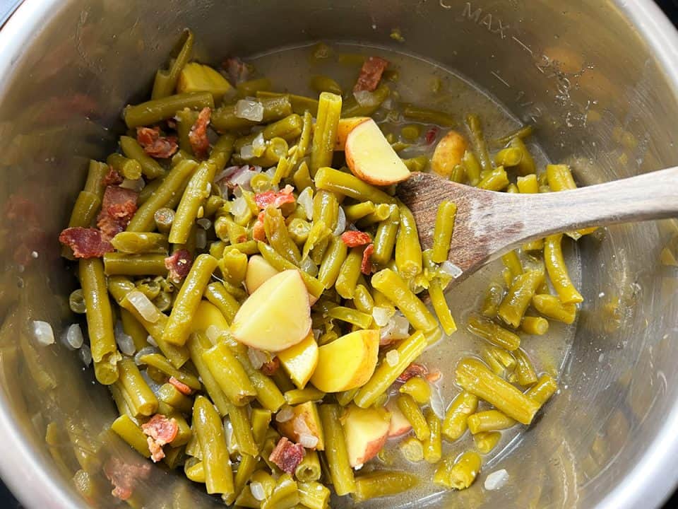 Green beans with bacon pieces, chopped potatoes, and dice onions being stirred in an Instant Pot.