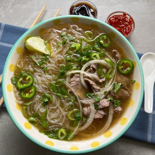 A bowl of finished beef pho with a side of chili sauce and a lime wedge, ready to eat.