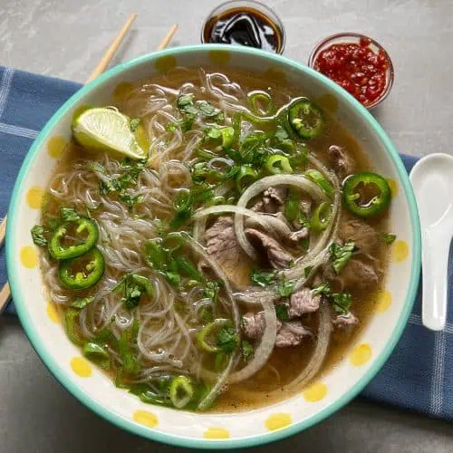 A bowl of finished beef pho with a side of chili sauce and a lime wedge, ready to eat.