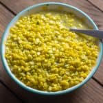 Instant Pot Creamed Corn in white bowl with teal rim and a spoon on wood table.