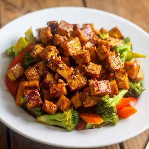 Tofu stir fry with vegetables in white bowl on wood table with chopsticks.