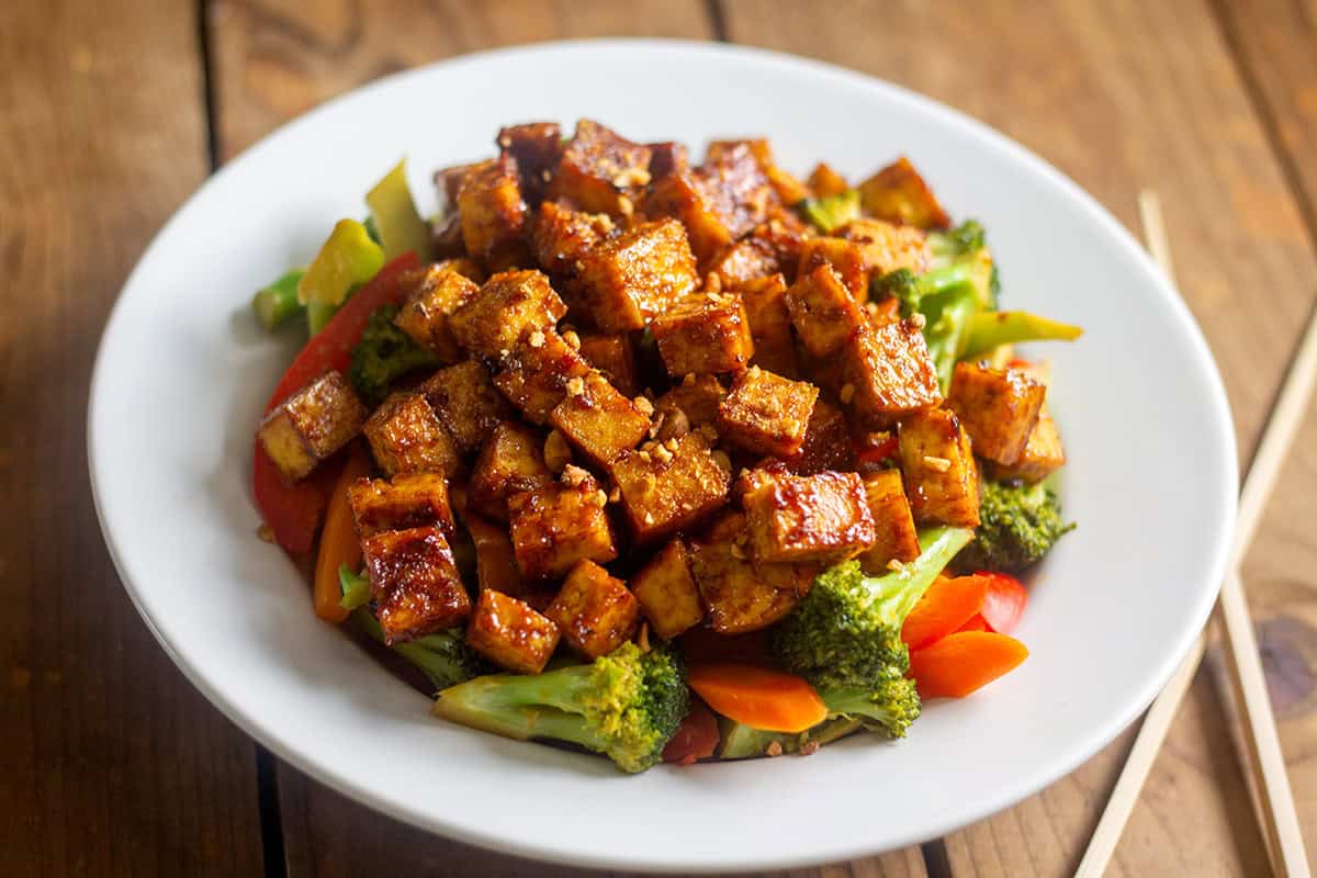 Tofu stir fry with vegetables in white bowl on wood table with chopsticks.