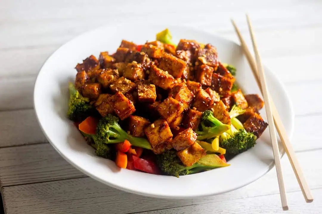 Tofu stir fry with vegetables in white bowl on white table with chopsticks.