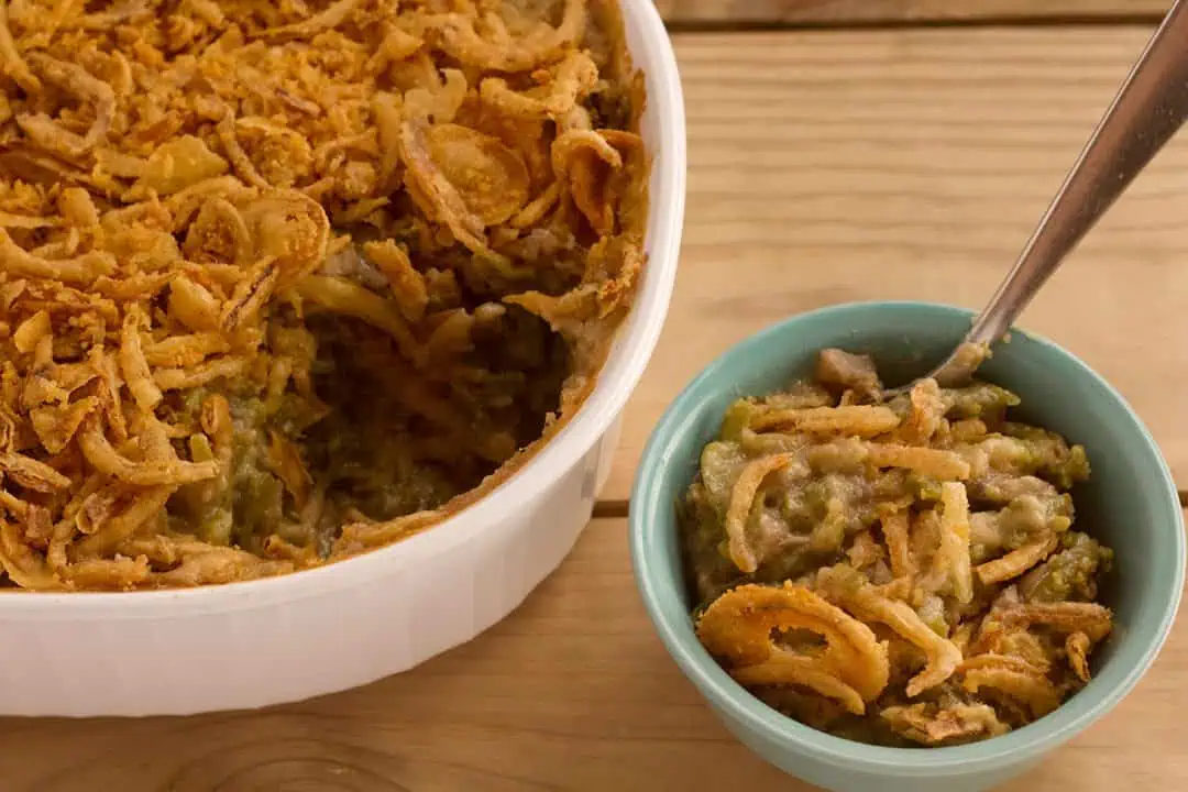 Two dishes of vegan green bean casserole on wood table with spoon.