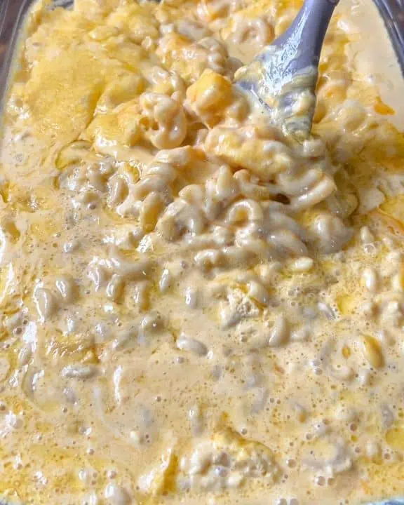 A casserole dish filled with macaroni, cubes of cheddar cheese, and egg custard, actively being stirred.