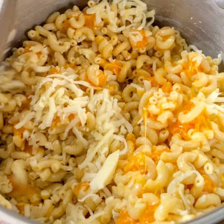 A pot filled with macaroni, small cubes of cheddar cheese, and shredded mozzarella, actively being stirred to melt the cheese.