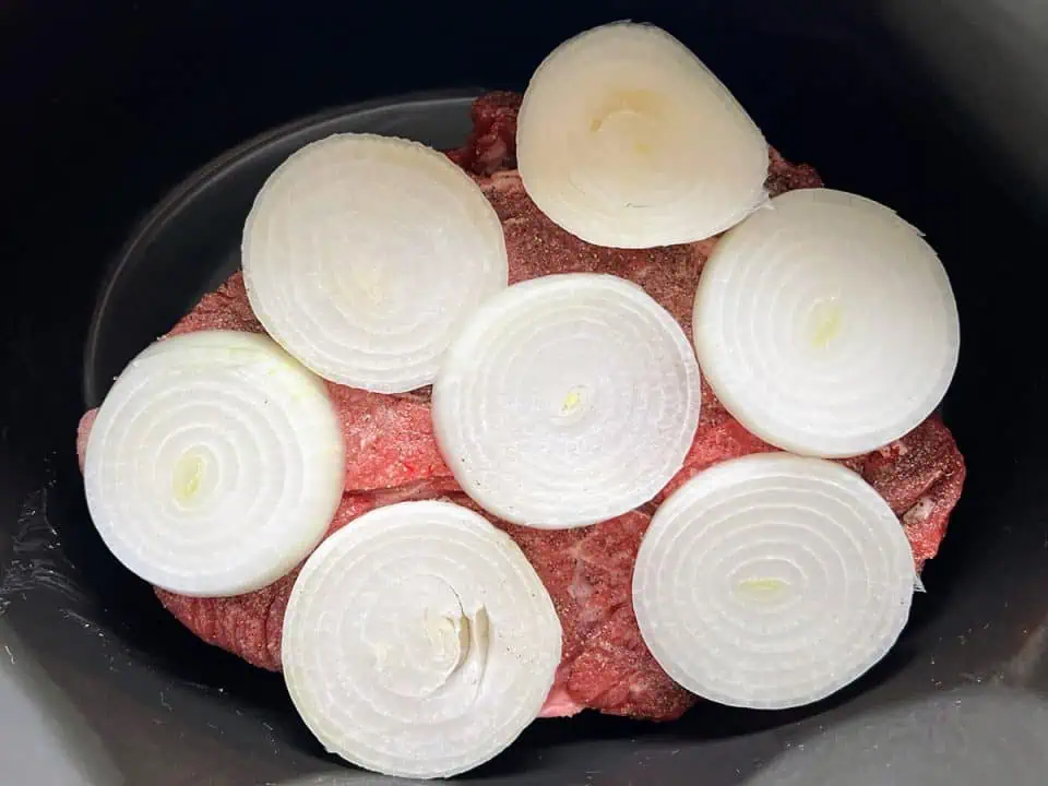 A Crock Pot filled with an uncooked pot roast topped with sliced onions.