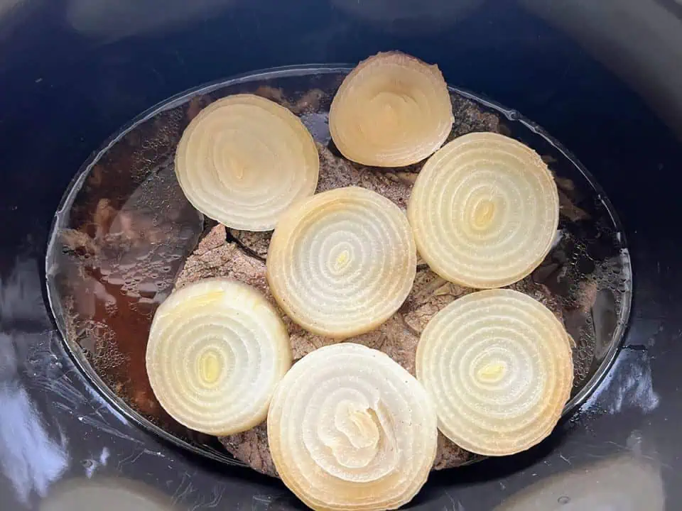 A Crock Pot filled with a cooked pot roast and au jus, topped with sliced onions.