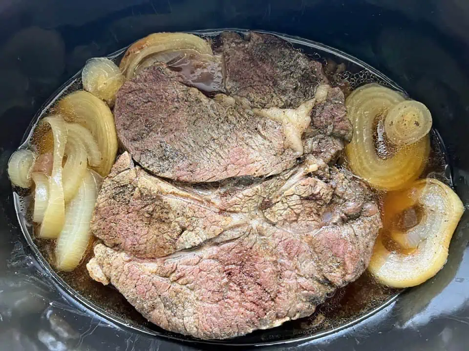 A Crock Pot filled with a cooked pot roast and au jus with sliced onions on the side.