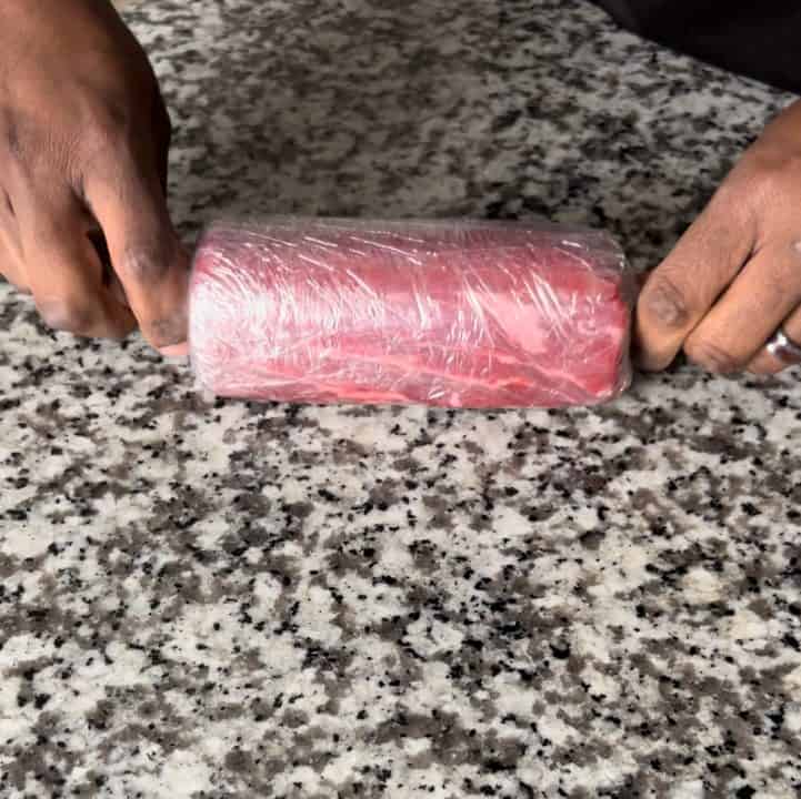 A person rolling center cut of beef tenderloin, using plastic wrap, into a perfect cylinder shape.