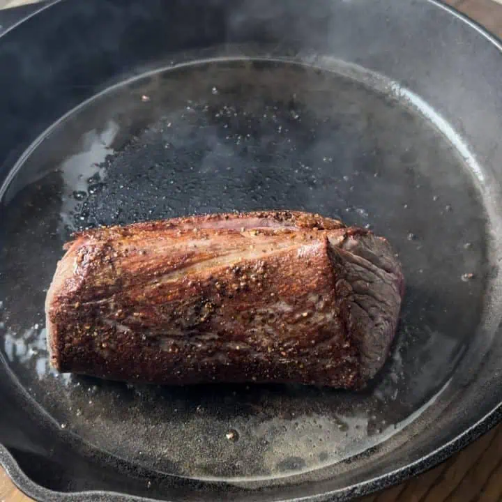 A chateaubriand cooks in a cast iron skillet, surrounded by steam.