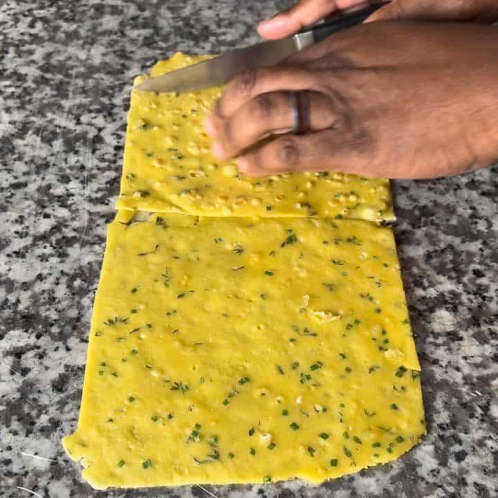 A person trims pieces of a yellow crepe with a knife on a granite surface.