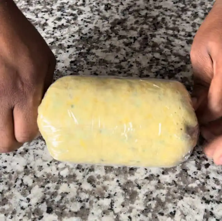 A person rolling a stuffed crepe, using plastic wrap, into a perfect cylinder shape.