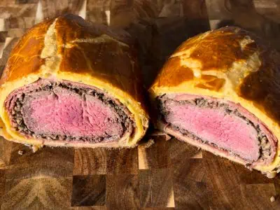 Beef Wellington on wood cutting board, sliced in half, showing the inside layers of filet mignon, mushrooms, prosciutto, crepe, and crispy puff pastry.