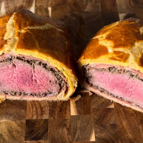 Beef Wellington on wood cutting board, sliced in half, showing the inside layers of filet mignon, mushrooms, prosciutto, crepe, and crispy puff pastry.