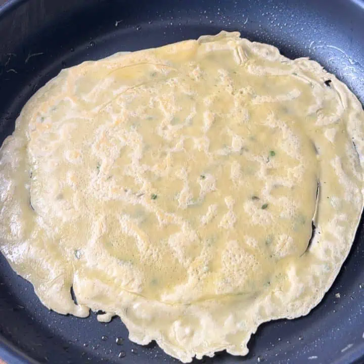 A thin chive and thyme crepe cooking in a nonstick pan.